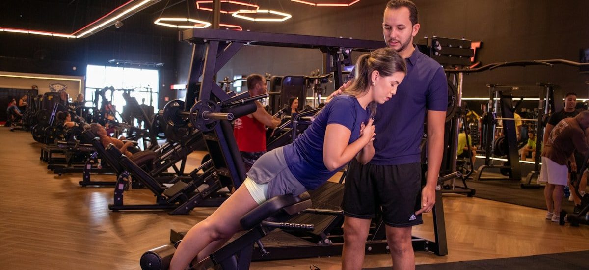 Mulher realizando exercício de agachamento em máquina de musculação com acompanhamento de personal trainer em academia moderna. A cena mostra treino de força com orientação profissional, foco na execução correta e segurança. Ambiente interno com equipamentos fitness, iluminação artificial e clima de disciplina e saúde.