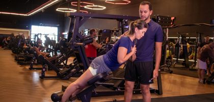 Mulher realizando exercício de agachamento em máquina de musculação com acompanhamento de personal trainer em academia moderna. A cena mostra treino de força com orientação profissional, foco na execução correta e segurança. Ambiente interno com equipamentos fitness, iluminação artificial e clima de disciplina e saúde.